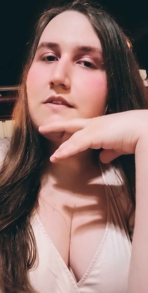 Tanya posing with their hand resting against their chin as they look towards the camera. They have on pale pink makeup and are wearing an elegant champagne colored dress.