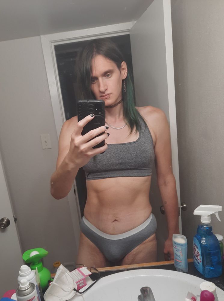 a white trans woman with black and green hair wearing a gray sports bra and gray panties