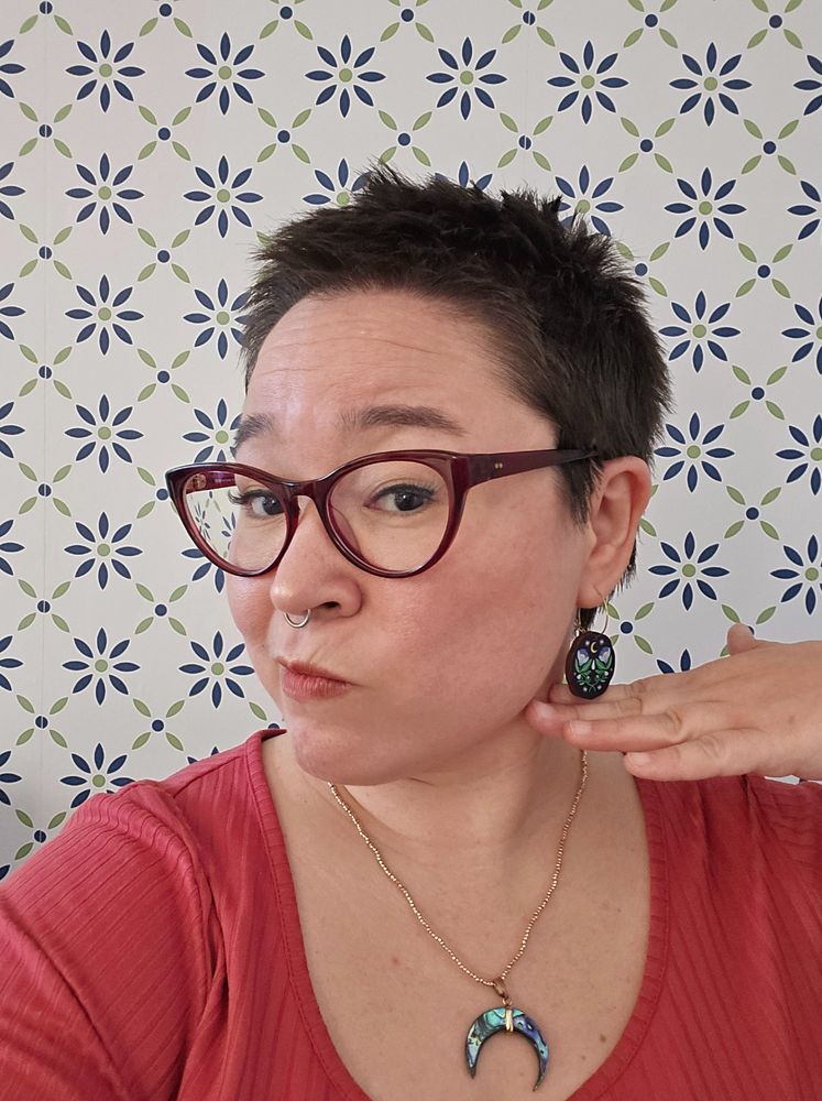 Woman standing in front of a wallpapered wall, showing off her new haircut and cool earrings. Nice, nice. 