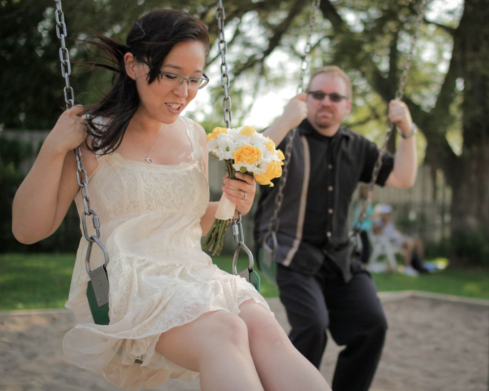 A man and a woman posing for wedding photos, this time on a swing set at a local park. The woman with dark hair and cream coloured lacy dress is holding a bouquet of white and yellow roses. The man is wearing a dark shirt and pants and his glasses have transitioned into sunglasses.