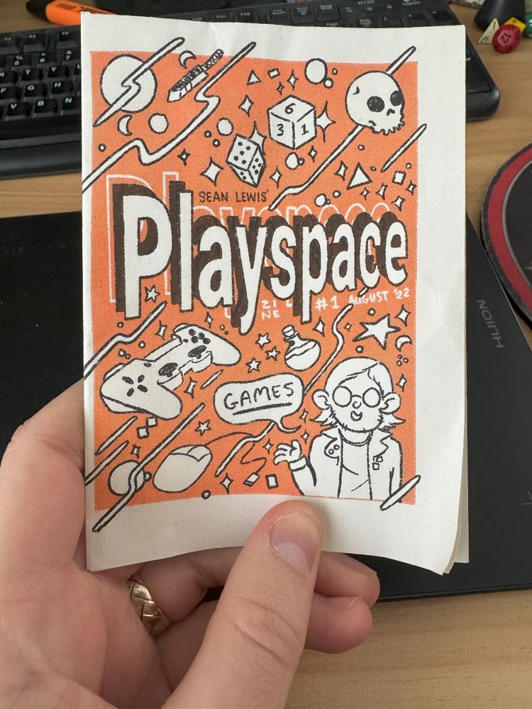 a photo of my zine PLAYSPACe, it features an orange cover with lots of cartoony gaming related things on it like dice, controllers, and particles, along with a cartoony self portrait of me with a word bubble saying "GAMES"