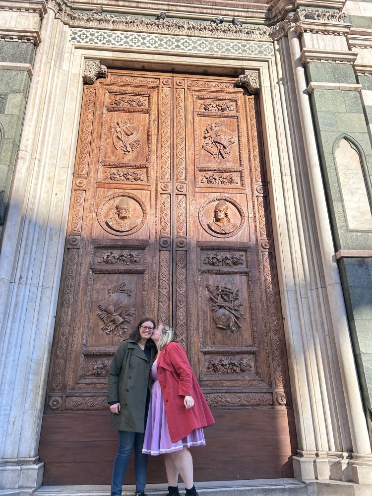 My wife and I, in front of the door at the Duomo in Florence. It’s 10am and bright outside. She’s wearing a green trench coat, jeans, and glasses. I’m wearing a purple swing dress with a red trench coat. I am kissing her cheek, and we are so happy.