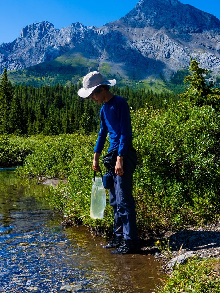 Picture of a young man gathering water from a creek with the Rockies in the background.