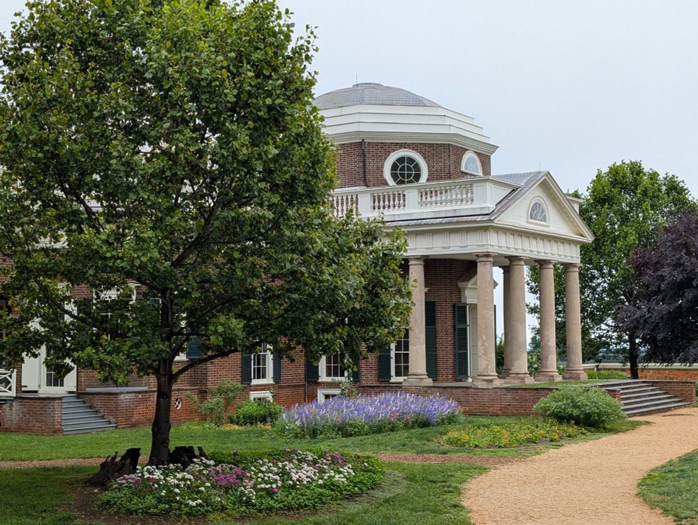 Brick domed building with pillars. A dirt path follows from foreground to its entrance. A tree sits in the foreground to the left. There are also flower gardens under the tree and beside the building.
