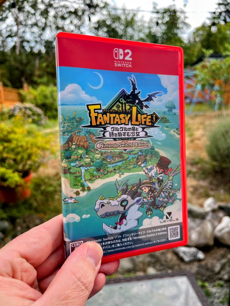 My hand holding a physical Japanese copy of Fantasy Life i for the Switch 2, showing off the front of the case, which features a cartoony illustration of three characters riding a dragon as it flies high over a fantasy island.