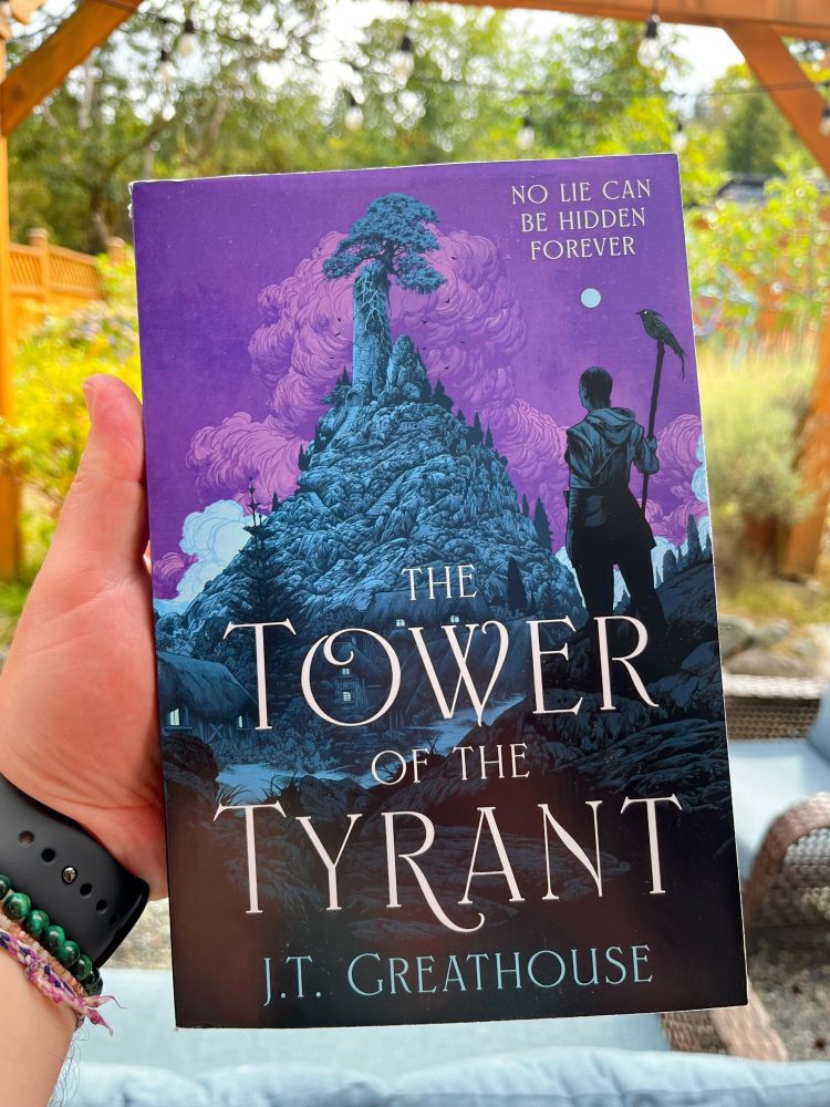 My hand holding a copy of “The Tower of the Tyrant” by JT Greathouse. A man overlooks a towering hill. Slate blue and purple predominate.