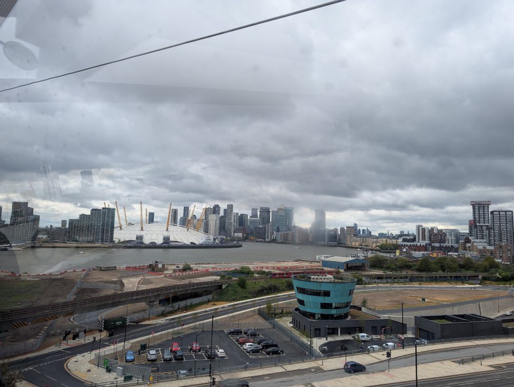 Image taken from inside a cable car crossing the Thames. It shows the 03 and Canary Wharf ahead. It's a very cloudy day so lots of low lying clouds amongst the buildings. 