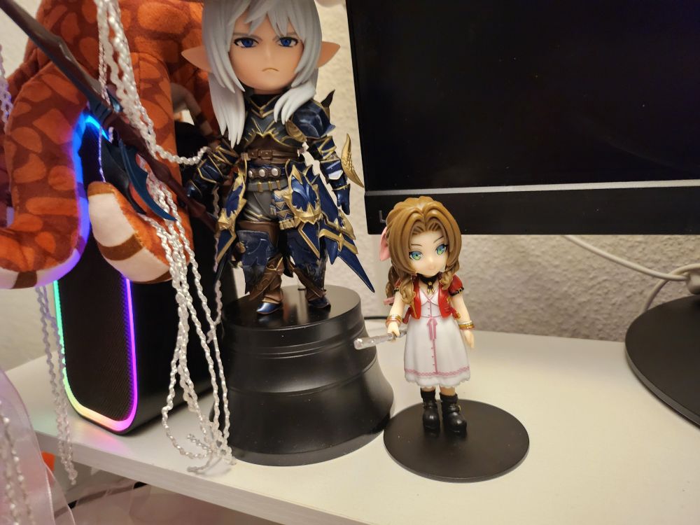 Close up of a desktop. There is a figure of Estinien from Final Fantasy XIV, and beside him is a figure of Aerith from Final Fantasy VII. Aerith appears to be about half the size of Estinien. In the background is the corner of a computer monitor and a computer speaker with an orange octopus plushie sitting on top.