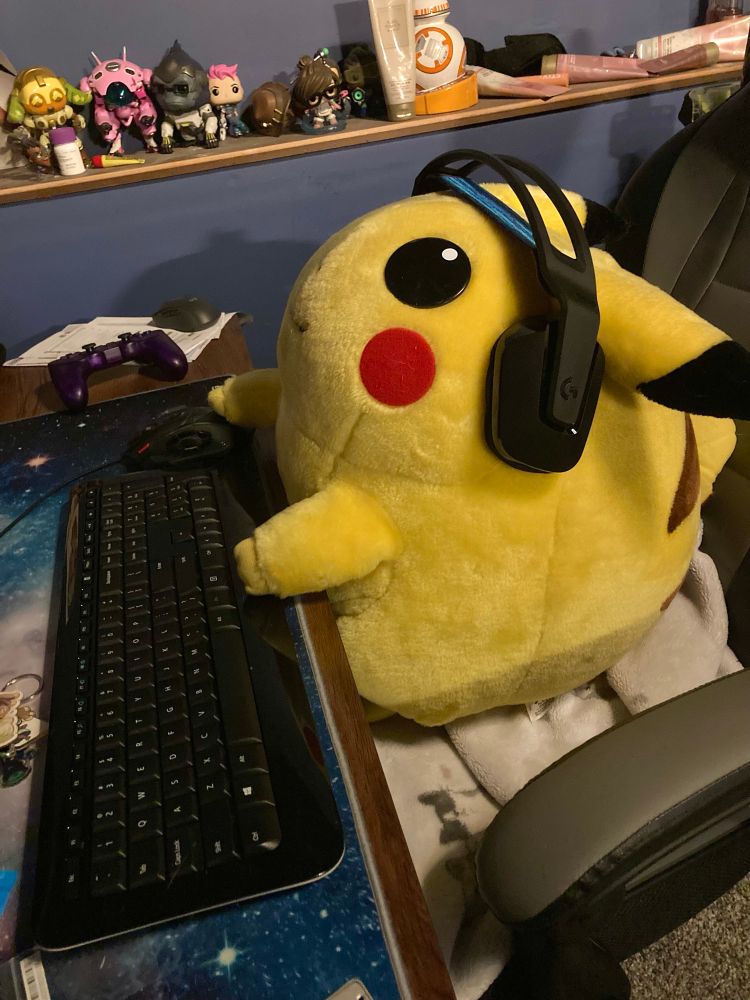 A fat pikachu plush sits at a computer desk with headphones on. The pikachu is playing Minecraft.
