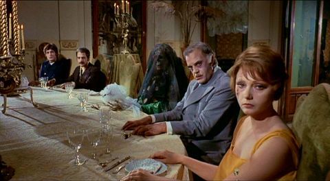 At a dinner table in a mansion, five corpse turn their heads and stare down the camera. One is a skeleton shrouded in a black veil. They have been positioned on one side of the table to resemble the Last Supper. 