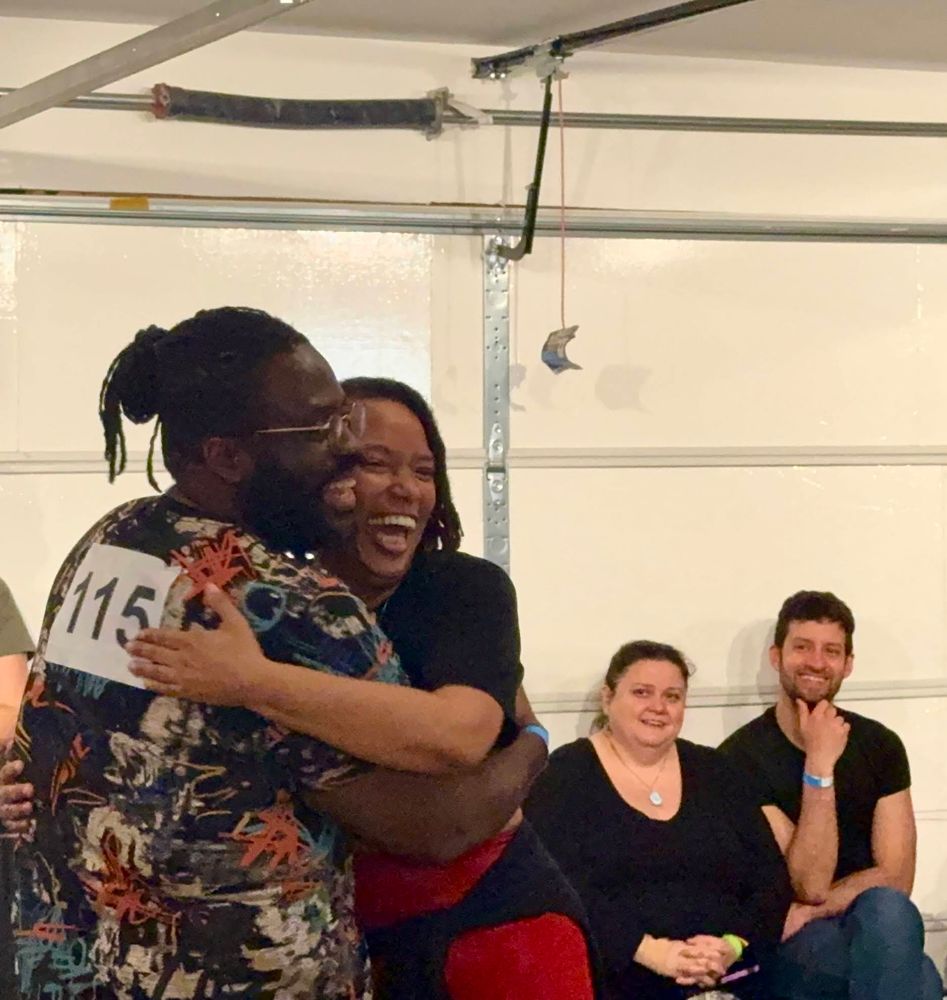 2 contestants sitting on a chair looking on 2 contestants hugging each other, the lead  with his back slightly turned to the camera wearing number 115 bib, both looking at the judges with the biggest smiles.