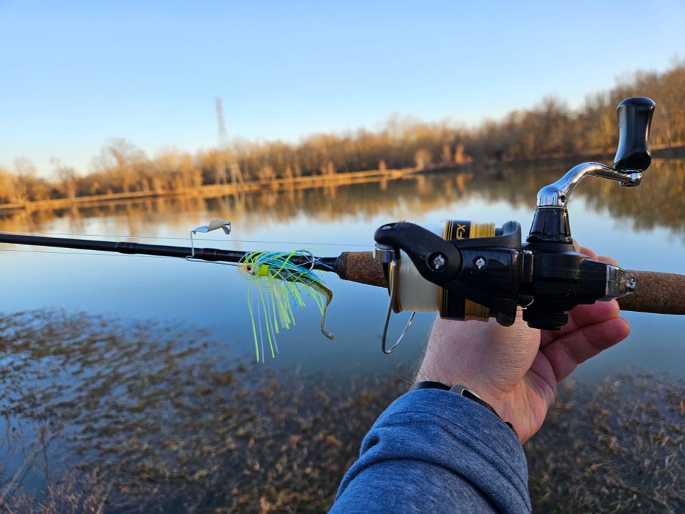 Arm holding a fishing pole with a spinning reel and a chartreuse and white buzz bait attached. The background is a lake in the late afternoon.