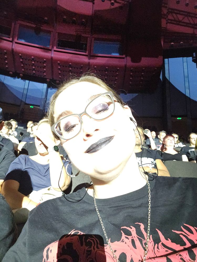 Konsertselfie av meg der hele ansiktet mitt er helt hvitt fordi lyset er kraftig. Jeg myser med øynene og smiler. Man ser at jeg har briller og mørk øyesminke og leppestift, og ellers ikke så mye detaljer på ansiktet. Dobbelthake, nesebor. Så vidt et øyebryn. Det er en person bak meg som skygger for ansiktet med hånda.
