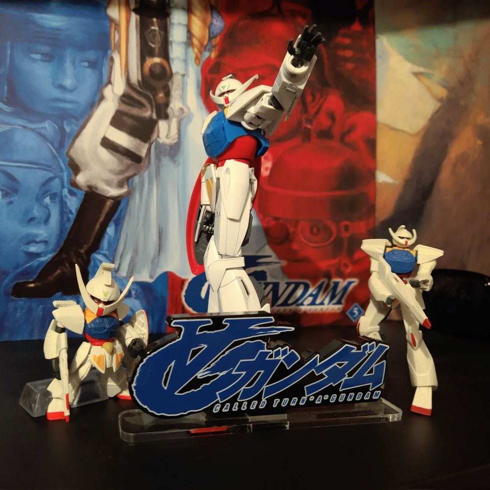 Photo of my 1/144 Turn A with gachapon Turn As on either side of it, and an acrylic logo stand in front of them. Behind them are laserdiscs volumes 5 and 2 of the series.