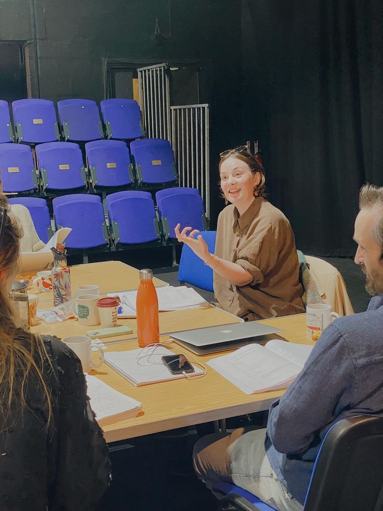 A woman animatedly speaks during a meeting in a dimly lit room, seated at a table with notebooks and drinks. Blue auditorium chairs are visible behind.