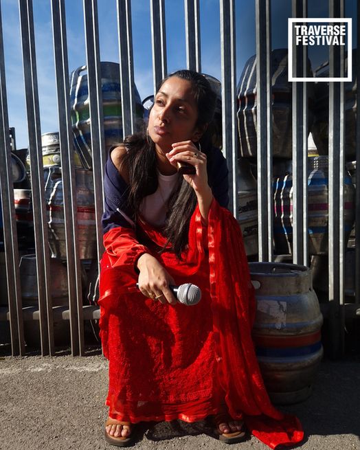 Woman in a vibrant red dress holds a microphone, seated in front of metal kegs with a thoughtful expression. "Traverse Festival" text in top right.