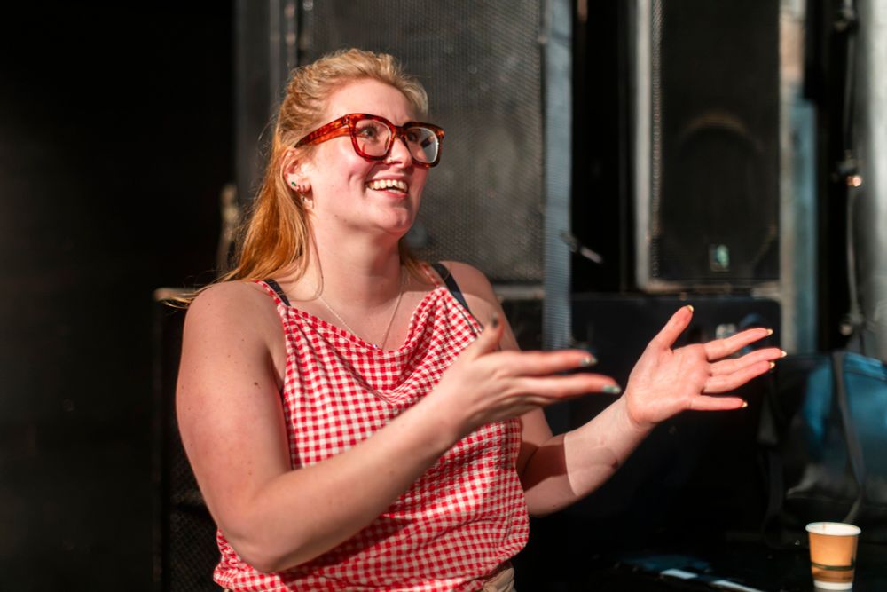 A person with long, light-colored hair and wearing large glasses is speaking expressively, smiling, and gesturing with both hands. They are wearing a red and white checkered sleeveless top. The background includes large black speaker equipment and a small paper cup on a surface nearby.