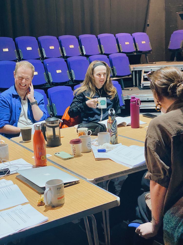 A group of people sit around a table cluttered with mugs, a French press, and papers, in a theatre with blue seats. They appear engaged and relaxed.