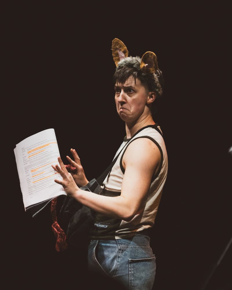 A person in a sleeveless shirt and jeans, wearing furry ears, holds a script marked with highlights, displaying a comedic, exaggerated expression on stage.
