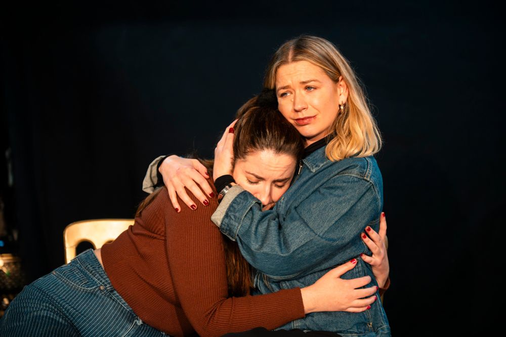 Two people sit closely together in a comforting embrace. One person, wearing a brown top and jeans, has their head resting on the other's chest, eyes closed. The other person, in a denim jacket, looks off into the distance with a concerned expression, gently cradling the other’s head. The background is dark, drawing focus to their emotional connection.