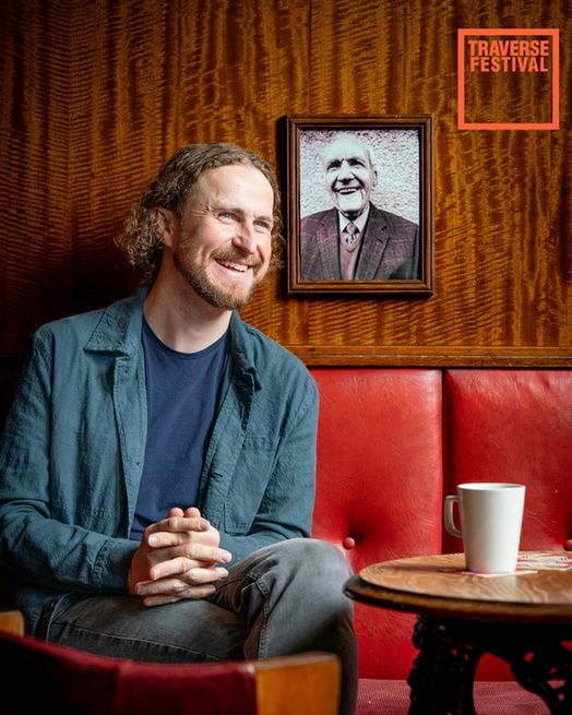 Smiling man sitting on a red couch in a cosy room, hands clasped, with a portrait of an elderly man on the wall. A white mug on a wooden table.