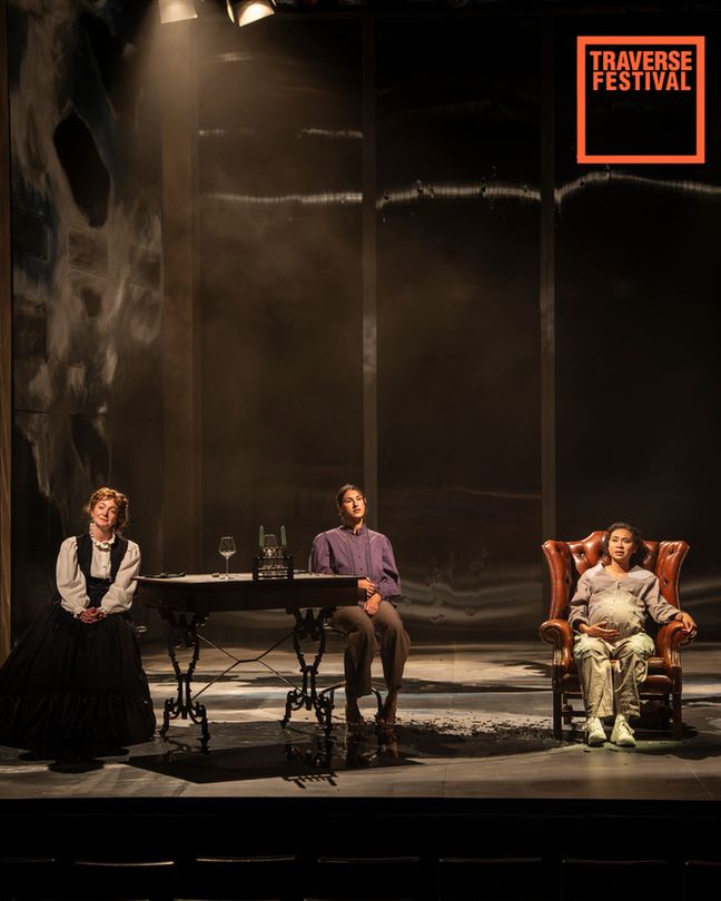 A dramatic stage scene shows three actors: one in a black dress, another seated at a table, and a third in an armchair. Atmosphere is tense, theatrical.