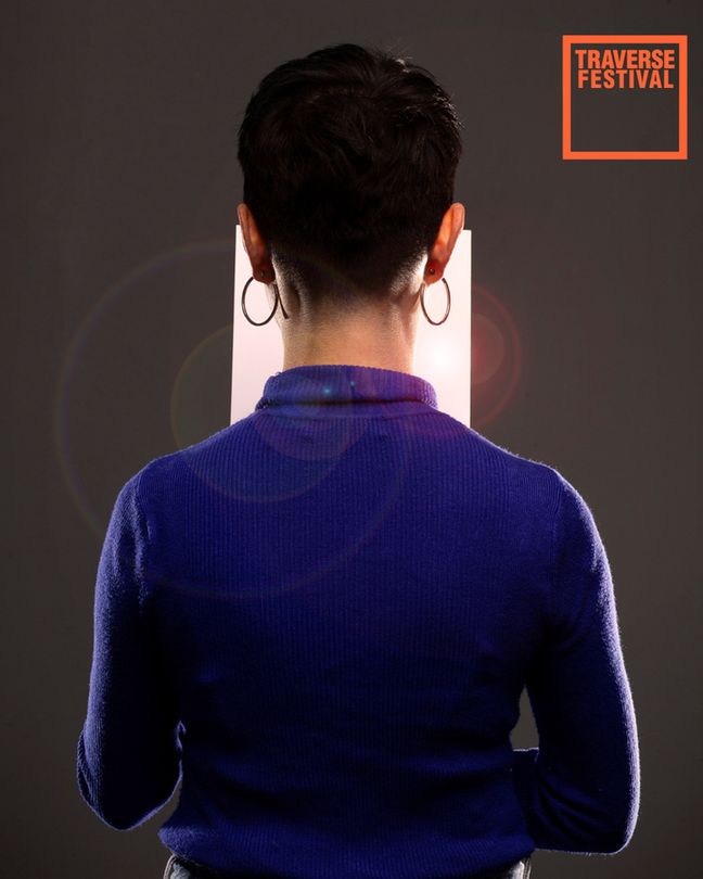 Back view of a person with short hair and hoop earrings, wearing a blue sweater. A bright rectangle lamp is in front of them. "Traverse Festival" text at the top.