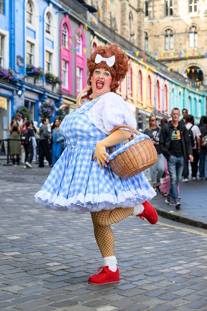 A lively performer in a theatrical costume poses playfully on a cobblestone street. The person wears a blue and white gingham dress with puffed sleeves, white lace trim, fishnet stockings, red shoes, and a large red wig topped with a white bow. They hold a wicker basket and strike a cheerful, exaggerated pose with one foot lifted. Behind them, a crowd of people walks along a colourful street with brightly painted buildings and hanging bunting, creating a festive atmosphere.