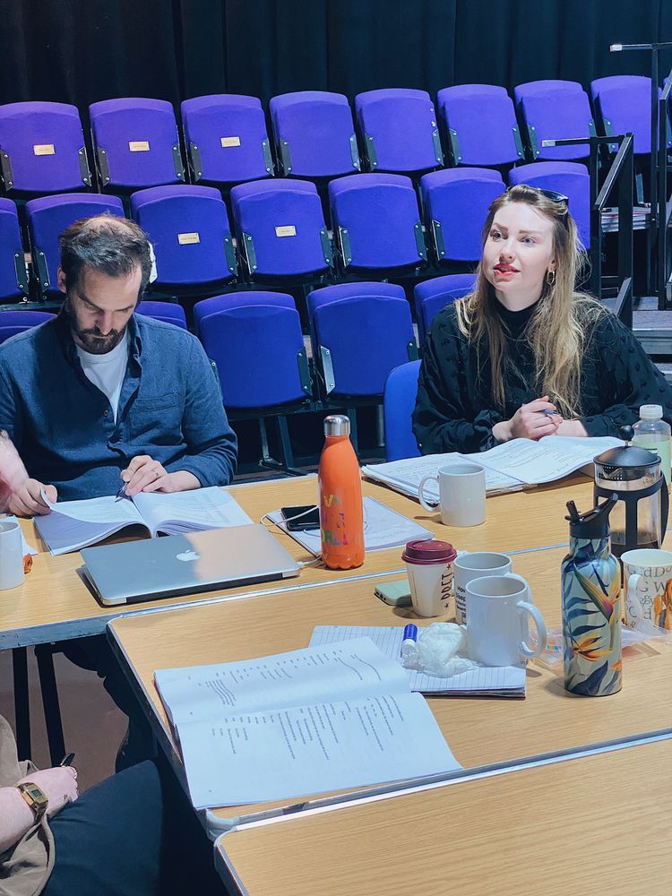 Two people sit at a table covered with scripts, laptops, and mugs in a brightly lit room. Behind them, rows of empty blue theatre seats.