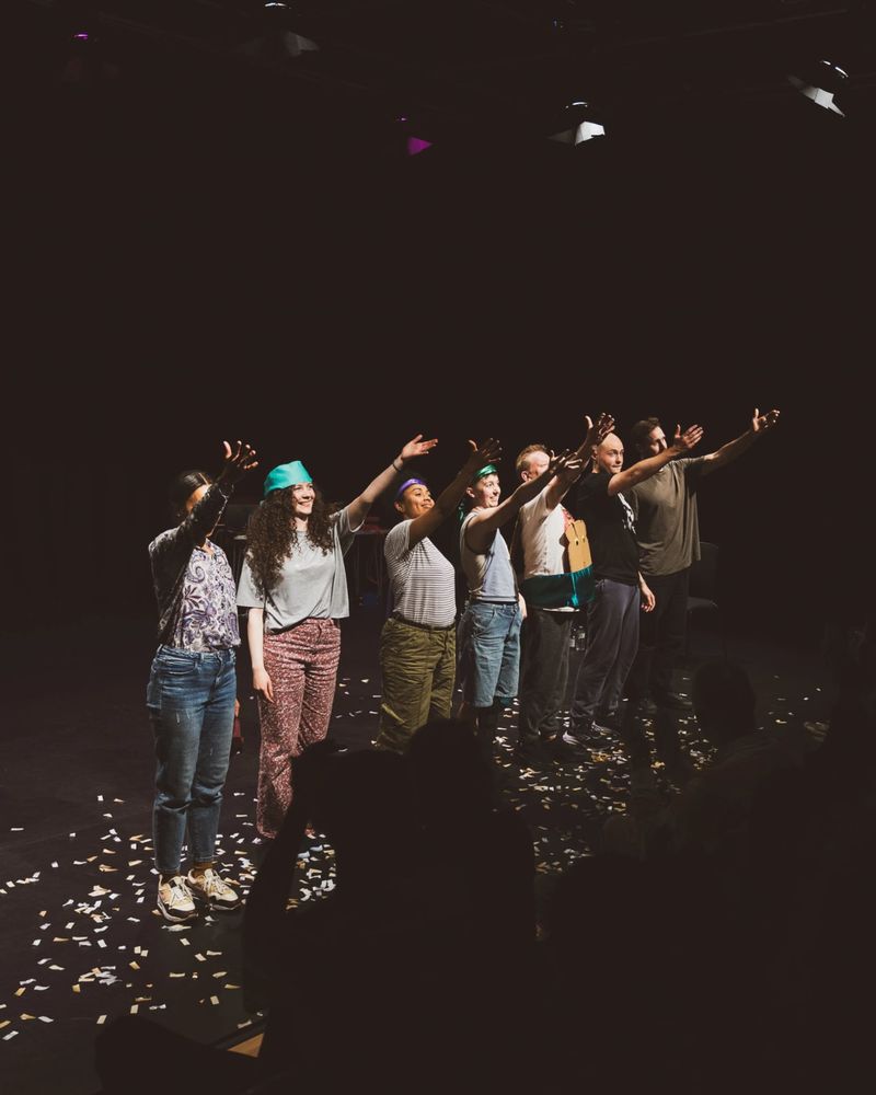  A diverse group of seven people stands on a dimly lit stage, smiling and waving at an audience. Confetti covers the floor, creating a celebratory atmosphere.