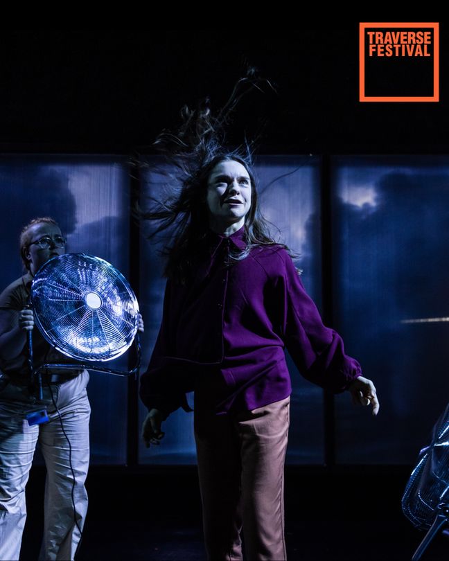 A woman stands dramatically with hair blown back by a fan held by another person. She’s wearing a purple blouse and peach pants. Background shows stormy skies. Text reads "Traverse Festival" in the top right corner.
