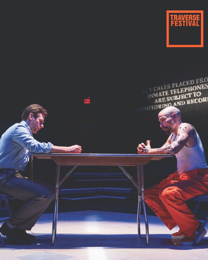 Two men facing each other across a metal table under low lights, creating a tense atmosphere. One wears casual office clothes, the other, tattoos and orange prison attire. The word "Traverse Festival" is displayed above.
