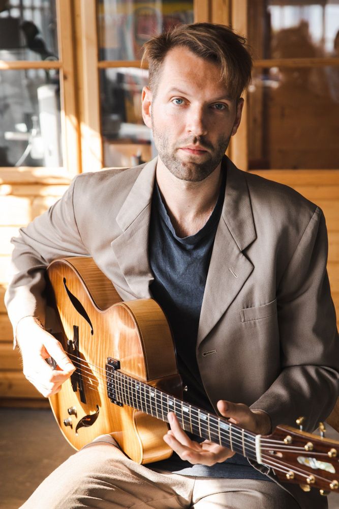 A man in a beige suit plays an acoustic guitar in warm light, sitting in a cosy wooden interior. His expression is thoughtful and focused.