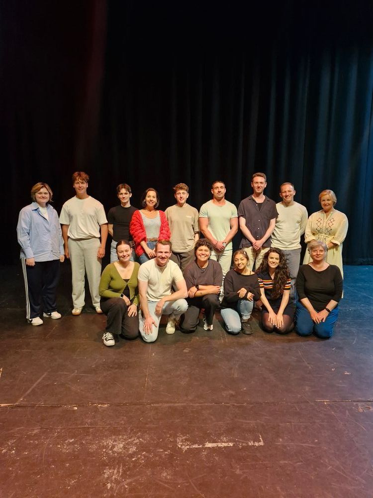 A group of fifteen people pose on a theatre stage, smiling warmly. They are arranged in two rows with a dark curtain backdrop.