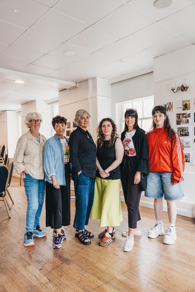 Six people standing together in a bright room with wooden floors. They wear casual, colourful outfits, conveying a friendly and relaxed atmosphere.