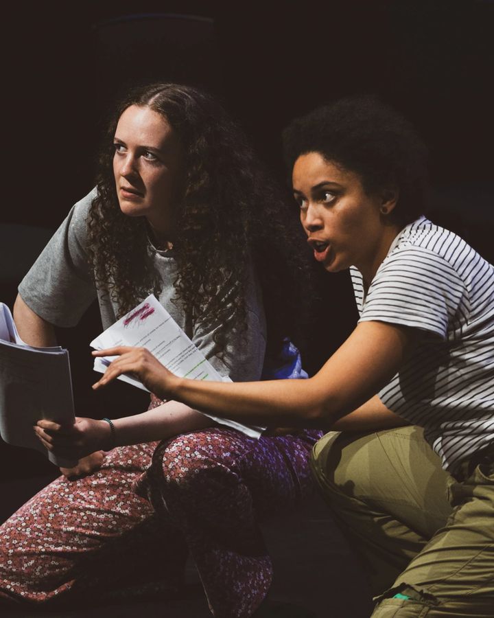 Two people kneel on a dimly lit stage, engrossed in rehearsal. One holds scripts and looks intently, while the other gestures expressively, conveying intensity.