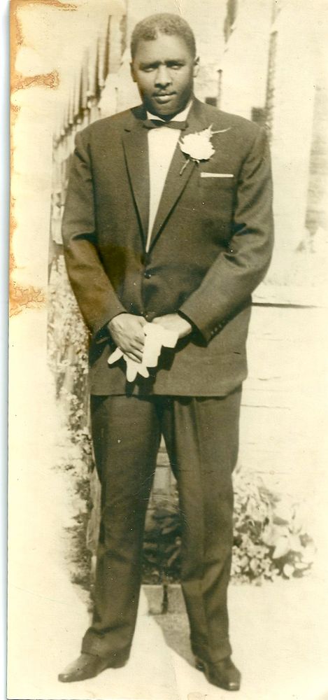 Tall black man wearing a dark suit with a white flower in the buttonhole and white gloves.