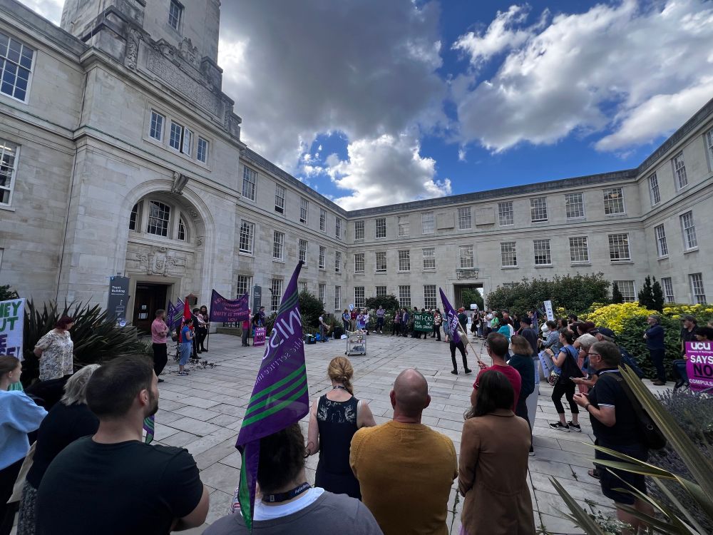 A wide angle shot of the same courtyard. Many people are standing around, waving flags and banners from different unions. In the background someone is giving a speech