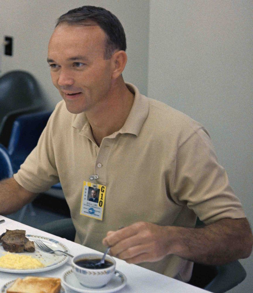Wearing a tan polo shirt, Michael Collins smiles while sitting at a table with a cup of coffee and a plate of steak and eggs in front of him.
