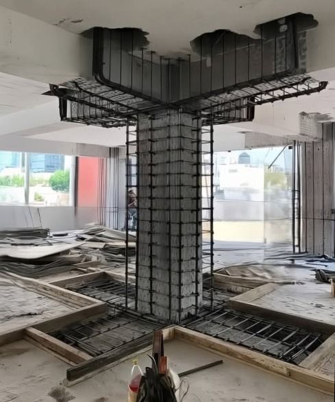 Structure en béton armé en forme de T en cours de construction dans un bâtiment, avec des barres d'acier visibles et matériaux environnants.