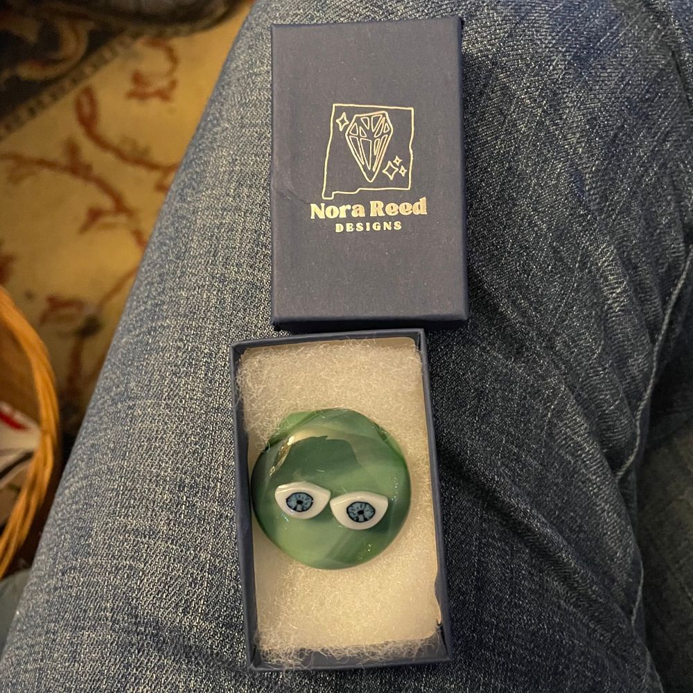 Balanced on a denim-wearing leg: an opened small dark blue box. The lid says “Nora Reed Designs” in silver. The bottom has a round glass blob in various shades of green with eyes.