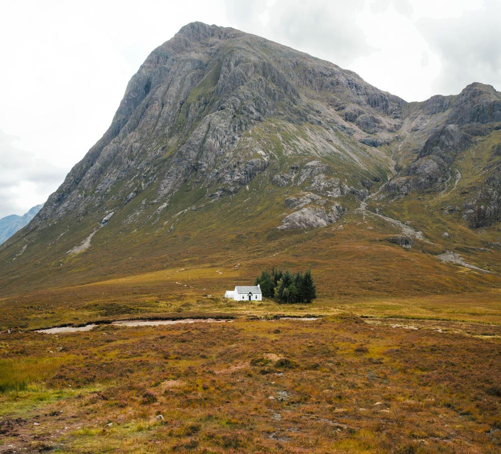 A view of the small white house on the way to Glencoe. The house sits before Buachaille Etive Mòr, a mountain in Scotland. The flora around the house is generally warm-colored, with a mild red tinge. As the elevation increases, the yellows and golds yield to greens, before it all abates into greys on the Munro. 