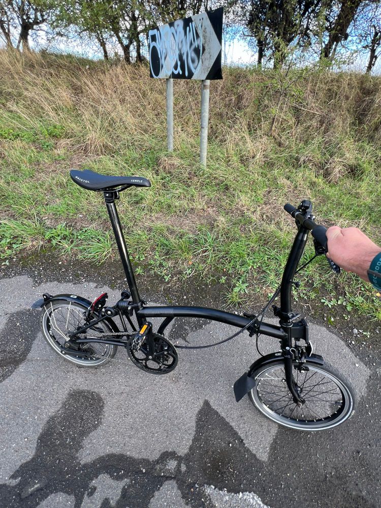 A black Brompton 2-speed folding bicycle held upright by the person taking the picture. The road under the bike had potholes and wet patches. In the background, a small grassy verge in which is planted a sign with chevrons that has been defaced. 