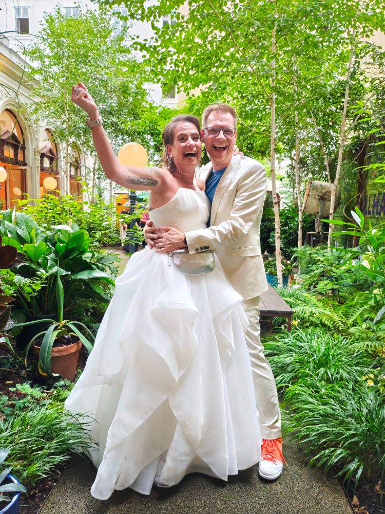 ALT-Text:
Ein fröhliches Hochzeitspaar steht lachend in einem grünen Innenhof mit vielen Pflanzen. Die Braut trägt ein weißes, voluminöses Hochzeitskleid, der Bräutigam einen cremefarbenen Anzug mit auffälligen orangefarbenen Sneakern. Beide wirken ausgelassen und glücklich.

