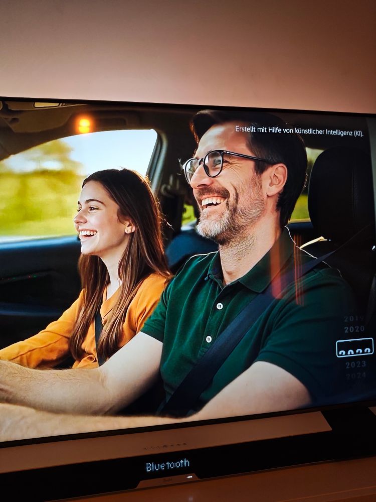 Ein Mann mit Brille und Bart sitzt lachend am Steuer eines Autos, neben ihm eine junge Frau, beide tragen Sicherheitsgurte. Oben rechts steht der Hinweis „Erstellt mit Hilfe von künstlicher Intelligenz (KI)“.
