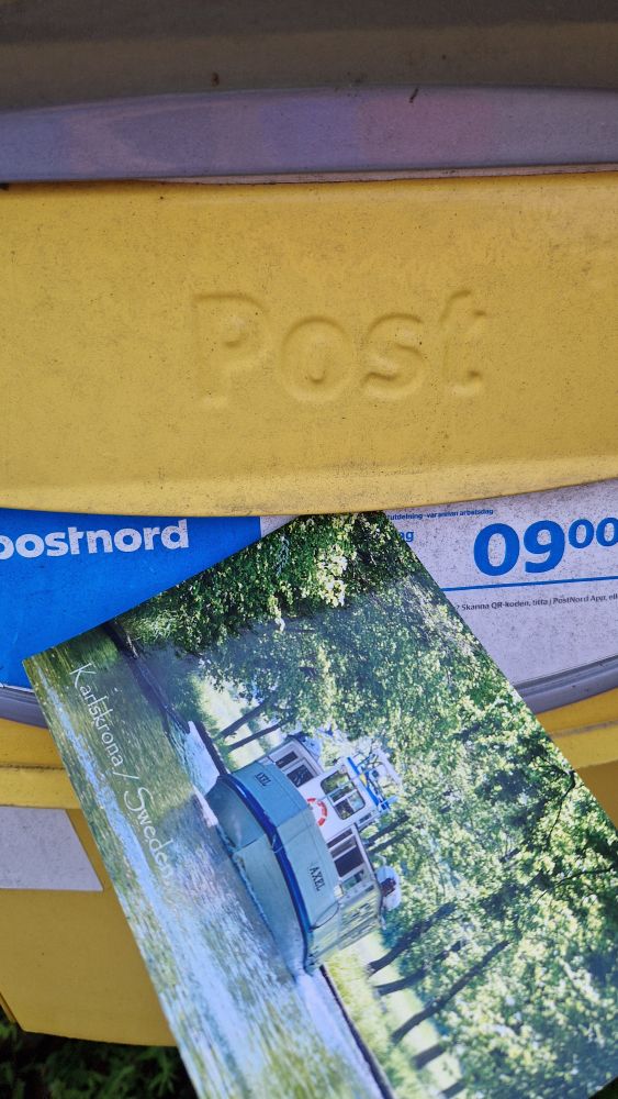 A yellow postbox with blue and white markings behind a close-up of a postcard with the photo of a steamboat plodding along in a tree-lined channel on it.