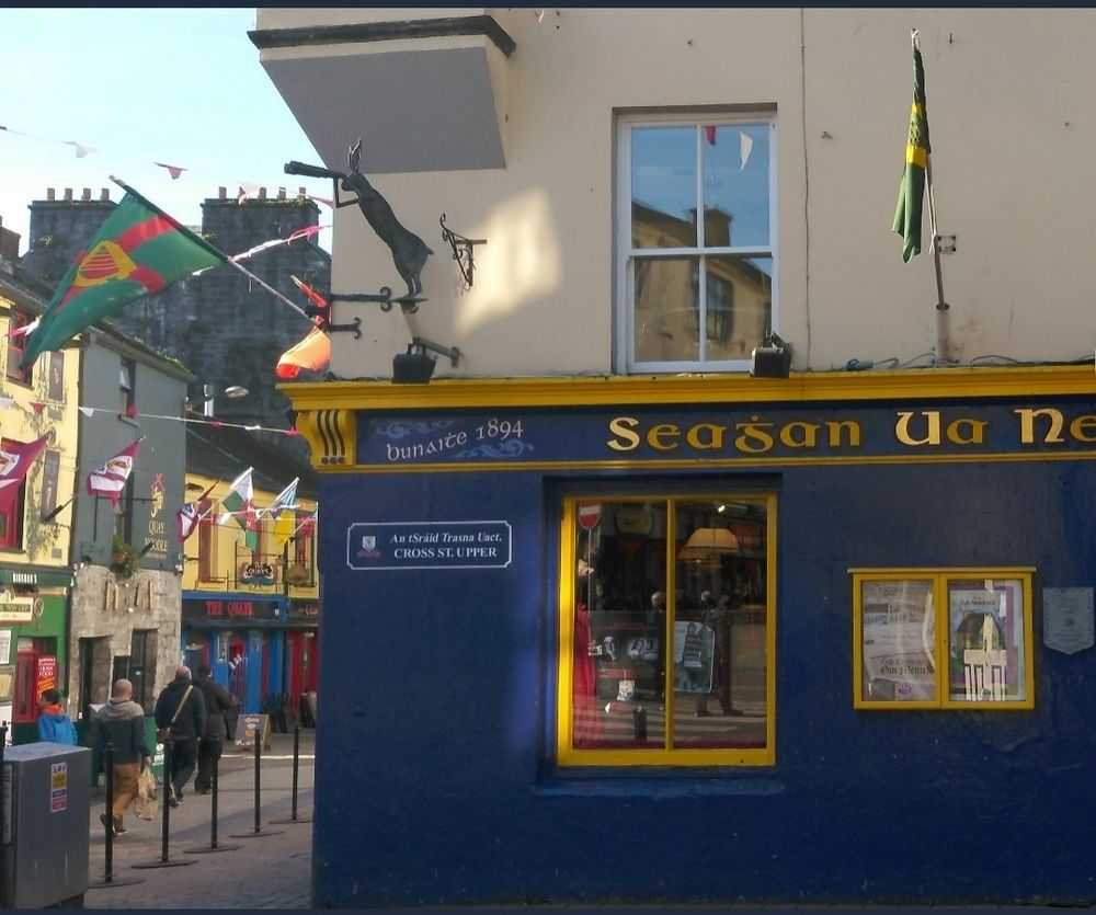 You see a pub in yellow and blue, to the right of the photo. It's Naughtons pub on Cross Street, Galway, Ireland. We see to the right, people walking down colourful Shop Street.