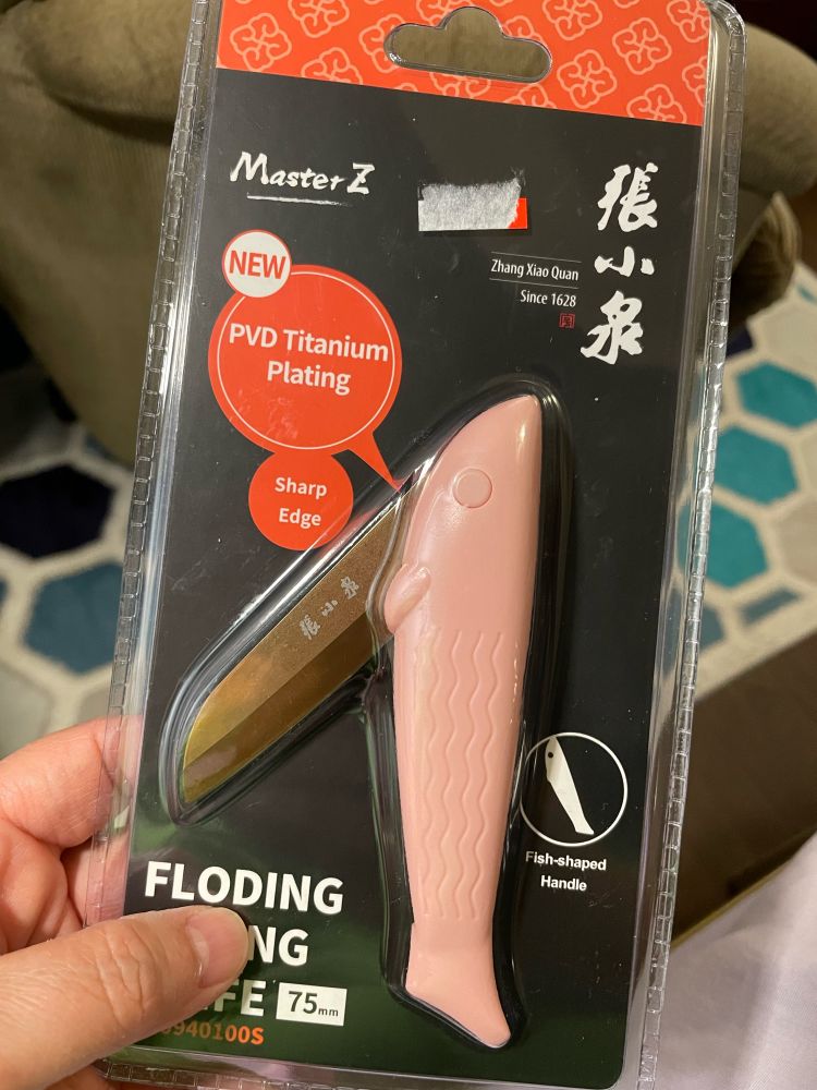 A hand holding the see-through plastic package containing a pink folding fish knife. The brand appears to be Master Z. “Folding,” notably, is misspelled “FLODING” (a thumb obscures the words “PARING KNIFE” under it). 