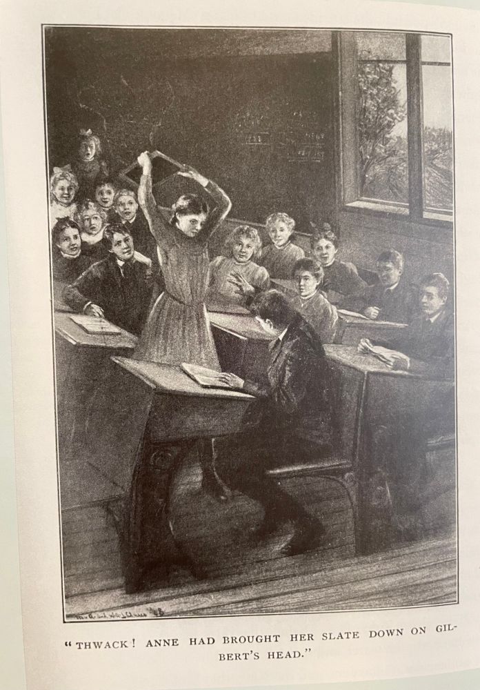 A black and white illustration by M.A. and W.A.J. Claus from the 1908 edition of Anne of Green Gables. We’re in a crowded classroom and Anne is standing over a seated Gilbert, whose futility trying to shield himself with his arm as Anne is about to bring down her slate, which she’s wielding in both hands high over her head, across HIS head. The other seated students watch the scene in anticipation from thier own desks. The caption under the illustration reads: “THACK! ANNE HAD BROUGHT HER SLATE DOWN ON GILBERT’S HEAD.”