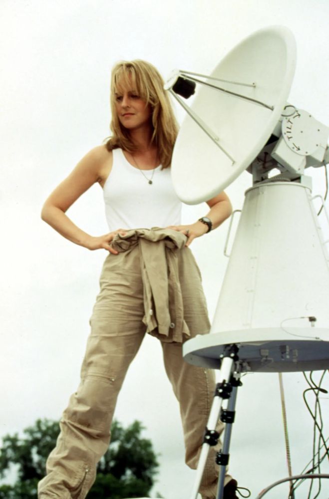 Helen Hunt as Jo from 1996’s Twister, a blonde woman with long hair.  She’s wearing a white tank top and khakis with a beige shirt tied around her waist, and is standing outside, on top of a van, hands on hips, next to a Doppler radar.